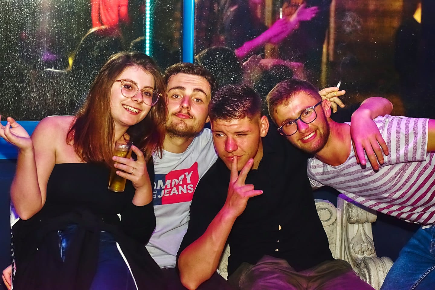 Vier lächelnde Freunde posieren in einem schwach beleuchteten Club mit Neonreflexionen.